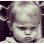 A young child with pigtails and a pouty expression looks directly at the camera, embodying a sulky demeanor in a black-and-white photograph.