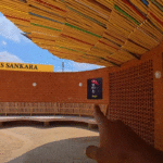 A person points toward the Thomas Sankara Memorial in Burkina Faso, with colorful roofing and a portrait of Sankara on the wall.