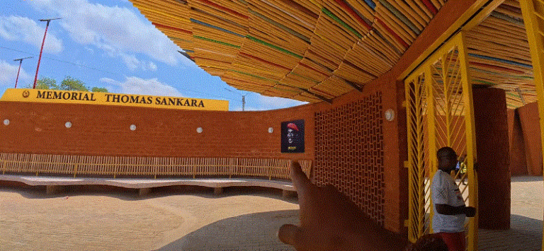 A person points toward the Thomas Sankara Memorial in Burkina Faso, with colorful roofing and a portrait of Sankara on the wall.